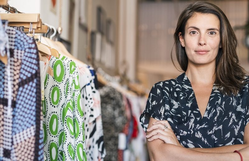 Portrait of female small shop owner with arms crossed in boutique. Mid adult woman is standing by clothing rack. Confident female is shopping in store.